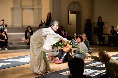 In the center of the sanctuary at St. Mark’s Church, Eiko Otake stands draped in white, she bends over reaching to hand a long stemmed flower to an audience member who is reaching forward to receive it. The audience sits all around the sanctuary, looking up at Eiko.