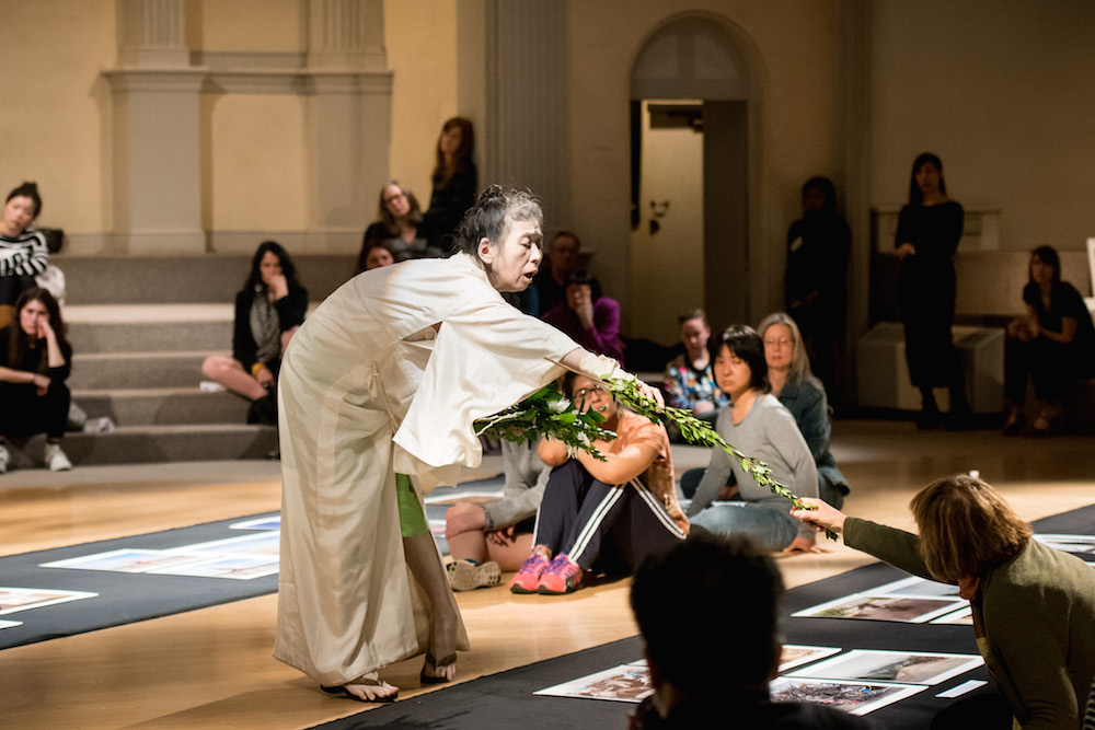 In the center of the sanctuary at St. Mark’s Church, Eiko Otake stands draped in white, she bends over reaching to hand a long stemmed flower to an audience member who is reaching forward to receive it. The audience sits all around the sanctuary, looking up at Eiko.