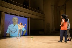 Hadar Ahuvia teaching a dance in front of a projected image of a man teaching a dance.