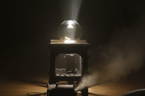 Set on a dark stage, an art object sits in the center. The object consists of a fog machine in front of a small table with a hole in the center where a stage light is set up underneath. Atop the table is a blown glass orb with a whiskey glass sitting at the base, the light shining a beam of white light that pierces through the glass and the dark empty space above.