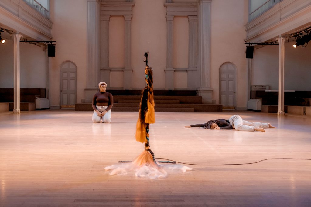 Two women in matching black turtlenecks and gray sweat pants are on the church sanctuary floor, one sitting on her knees upright, and one is lying on her belly curling in on the side of her body that faces the camera. A microphone stand, dressed in many thick brown, orange and white braids with green ribbons woven through, is foregrounded in the center.