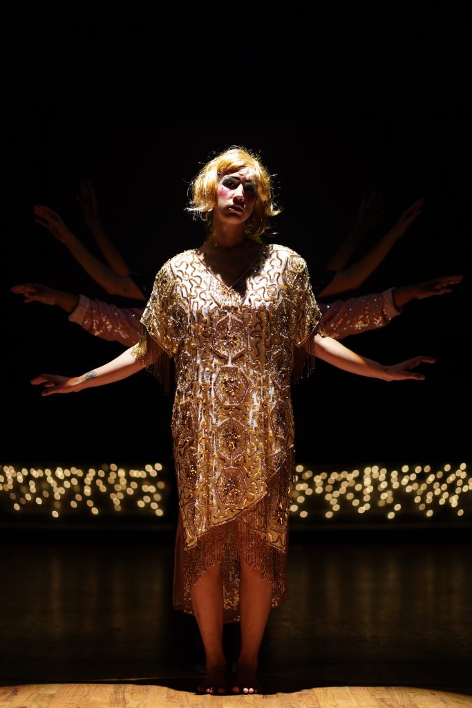 Dancer standing in parallel with hands held out to side, palms down, wearing gold sequins dress and gold wig.