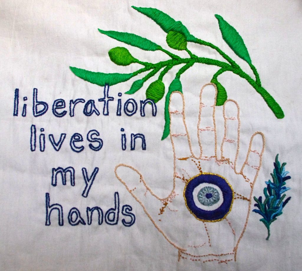 Royal blue, lowercase text that reads "liberation lives in my hands" is embroidered onto cream colored fabric. To the right of the text is an upright open hand embroidered in different shades of brown and pink. A large blue eye is stitched into the palm of the hand, known as mati, nazar and/or Khamsa. To the right of the hand is a small rosemary branch embroidered in greens and blues. A bright green olive branch is stitched into an arch above the text, Khamsa and rosemary.
