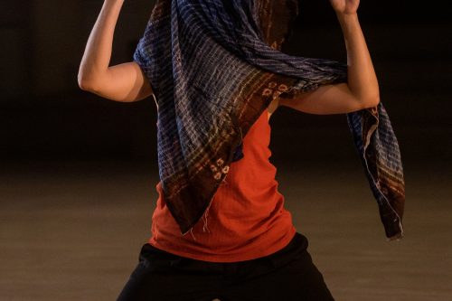 Woman on her knees with both arms bent at her side with her index fingers pointing up, she has a large blue scarf wrapped around her head, covering her whole head.