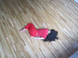 A photo taken from above, we look down on a woman dressed in all red, curled laying on a sandy, blonde, hard-wood floor.