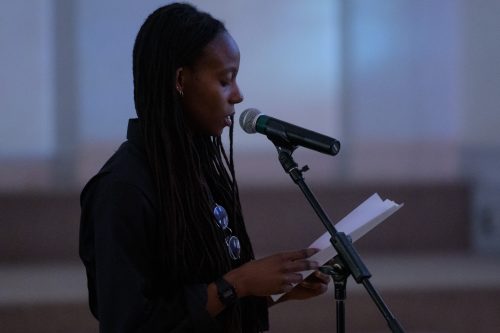 Young woman speaking into a standing mic, eyes cast downward looking at the notebook she cradles h=in her hands. The lighting is low and soft and blue.