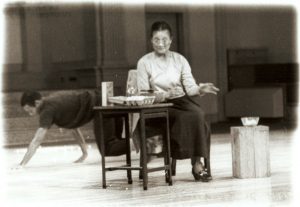 A faded black and white photo with Soft edges. In the Sanctuary stage of the St. Mark's Church, an older woman sits at a table dying eggs as a young man crawls on all fours behind her.