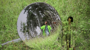 A projection of tall green grasses projected onto Eiko reaches an arm towards the images of a circular convex mirror that reflects the image of Joan in a neon green coat, orange hat, and dark sunglasses, standing in the grassy fields, the woods behind her.