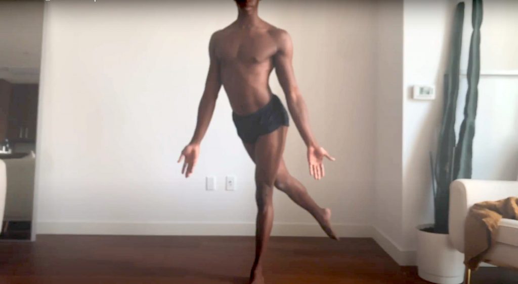 In a room filled with natural light, a dancer, Quenton Stuckey stands in front of a white wall and beside a cactus plant and sofa chair. Quenton stands on one leg with the other held in a pointed line, crossed, behind the standing leg. Hanging from a bare chest, Quenton's arms are held low, with open palms facing forward. Due to the frame of the photo, we can only see the chin and mouth of Quenton's face.