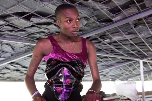 Yves turning hovering over her dj controller setup in a brightly lit industrial setting. She is wearing a highly textured corset top with purple velvet and slick black. She is wearing thin metal chains and rings, looking up as her hands are busy at work.