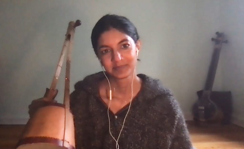 Samita smiles while looking into the camera and holding a small wooden string instrument in one hand. She wears a cozy grey sweater and white earphones. Another string instrument sits in the background.