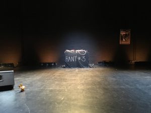 In a dark black box stage sits a small bunny toy next to an amp, and in the back center is a spotlighted soundboard with many wires hanging off of it, and on the front are the words, RANT #3 written in white chalk. On the wall behind is a framed picture.