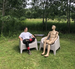 two seated bodies- one human one mannequin, in chairs with a table in a field