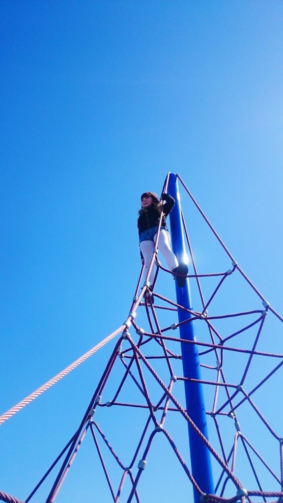 Low Angle View Of child on top of a web-like rope pyramid against a clear blue sky.