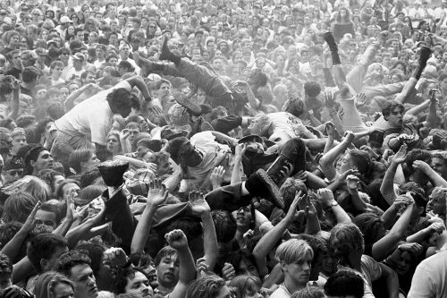 In black and white, a tightly packed crowd at a concert. Some are crowd surfing or climbing over others, several pairs of legs sticking up out of the crowd.