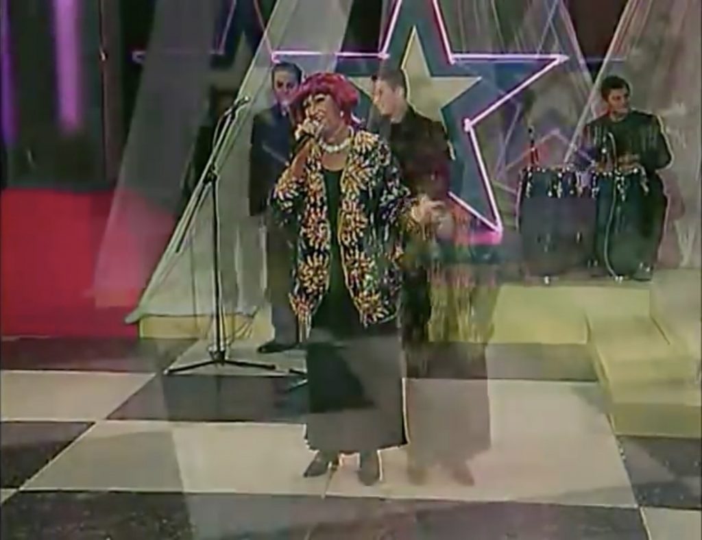 A freeze frame from a video of a live performance by singer Celia Cruz and band. The stage is decorated in neon purple stars and Ms. Cruz wears an elaborately adorned black and gold costume. Her hair is magenta