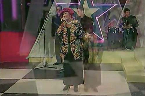 A freeze frame from a video of a live performance by singer Celia Cruz and band. The stage is decorated in neon purple stars and Ms. Cruz wears an elaborately adorned black and gold costume. Her hair is magenta