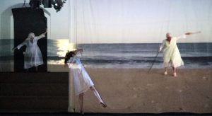 A grainy projection of a person standing on a beach wearing a white dress. They carry a stick and lift their other arm to shoulder height. In front of the projection, a person leans against the wall, standing on their toes. Another person in a white dress stands on large stairs adjacent to the wall. They look over at the projection, mirroring the projected person's body shape. The image of the beach projects onto both of their white dresses.
