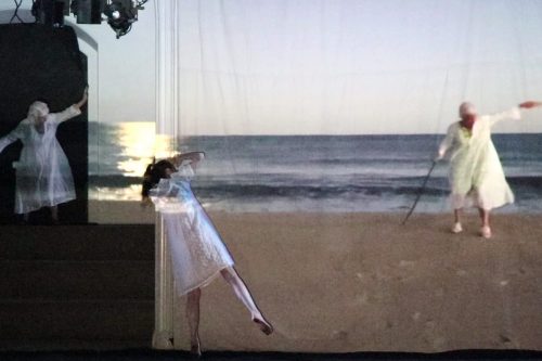 A grainy projection of a person standing on a beach wearing a white dress. They carry a stick and lift their other arm to shoulder height. In front of the projection, a person leans against the wall, standing on their toes. Another person in a white dress stands on large stairs adjacent to the wall. They look over at the projection, mirroring the projected person's body shape. The image of the beach projects onto both of their white dresses.