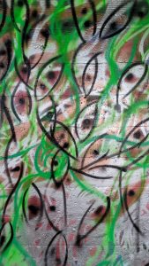 A spray painted abstract design. Many eye-like or fish-like shapes superimposed over each other. Colors are white, black, green, and brown.