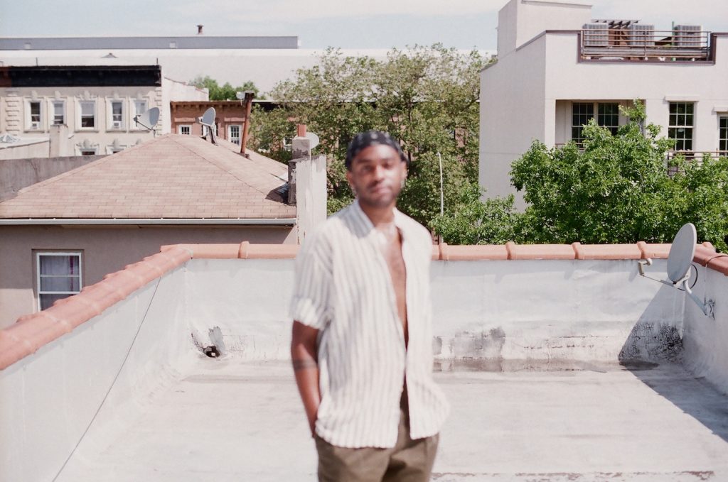 Jordan stands in a button down and bandana on what appears to be a rooftop. They are blurry and out of focus.