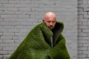 Christopher hugs a coat made of grass and conjures up the image of a raging mountain. His facial expression is intense and his forehead is sweating.