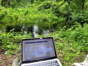 In front o a lush green landscape with a stream at its center, a sliver mac laptop open to a word doc.