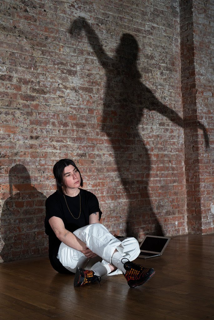 Gillian sitting in front of a brick wall watching rehearsal with a large shadow of a dancer on the wall next to her.