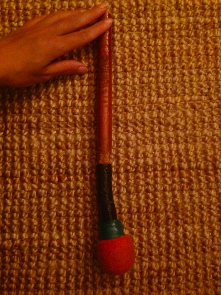 Mallet. A brown hand rests on a wooden mallet that has been wrapped in green and black tape. The soft felt tip of the mallet is bright red.