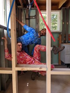 ​Rashaun and Silas are seen from behind vertical wooden beams for a wall that is not yet sheetrocked. The room they are in looks like it is mid-construction. Lots of exposed beams and pipes. They both wear colorful, patterned jumpsuits.