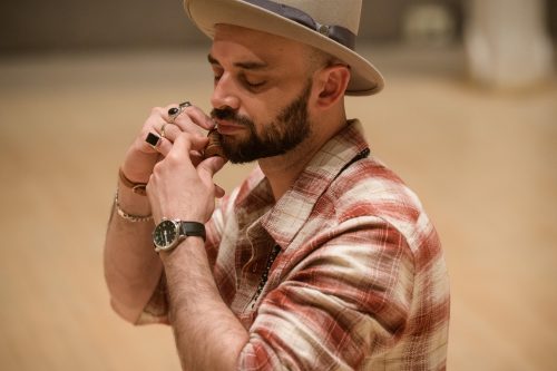 Christopher is in a tan hat and plaid shirt. He wears a watch and bracelets around his wrists and silver and black rings on every finger of his right hand. He holds an object against his bearded chin, with closed eyes. This object is an indigenous object made of mud from his birthplace of Costa Rica.