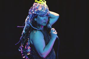 An Asian American woman, in a black sparkle dress, is on stage in purple/blue lights. She looks out at an audience mid-sentence, while holding a mircophone and wearing a headdress made of aluminum can ribbon tendrils, flouresecent green tentacles, and reclaimed plastics. Her makeup looks like black branches, tentacles or snakes growing from the corners of her eyes.