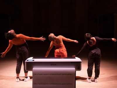 Three Black femme dancers with their backs turned, perform a synchronized and wide wingspan gesture, looking up over slightly lifted right shoulders. Infront of the dancers is a large industrial printer that has a long stream of paper extending out onto the blond wood floor of a stage, with lines of printed text covering the enormous page that reads: “OK so I found a squirrel in my backyard found a squirrel yeah there, OK so I found a squirrel in my backyard found a squirrel yeah there”