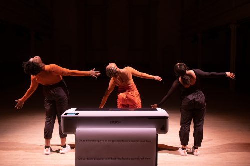 Three Black femme dancers with their backs turned, perform a synchronized and wide wingspan gesture, looking up over slightly lifted right shoulders. Infront of the dancers is a large industrial printer that has a long stream of paper extending out onto the blond wood floor of a stage, with lines of printed text covering the enormous page that reads: “OK so I found a squirrel in my backyard found a squirrel yeah there, OK so I found a squirrel in my backyard found a squirrel yeah there”