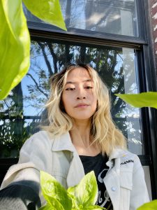 A selfie of Yo-Yo, a Taiwanese-American femme, sitting outside on her fire escape. It’s a sunny day, her hair is blonde and slightly wavy, she gazes serenely at the camera. Basil leaves frame the photo. Behind her is a window reflection of a tree and blue sky.