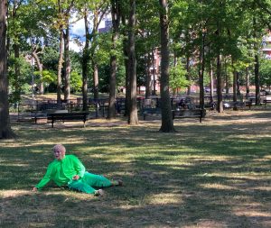 The artist with a bleached short hair and a white and multi-colored painted face in a green costume lying halfway down in a park.