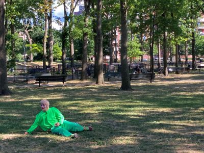 The artist with a bleached short hair and a white and multi-colored painted face in a green costume lying halfway down in a park.