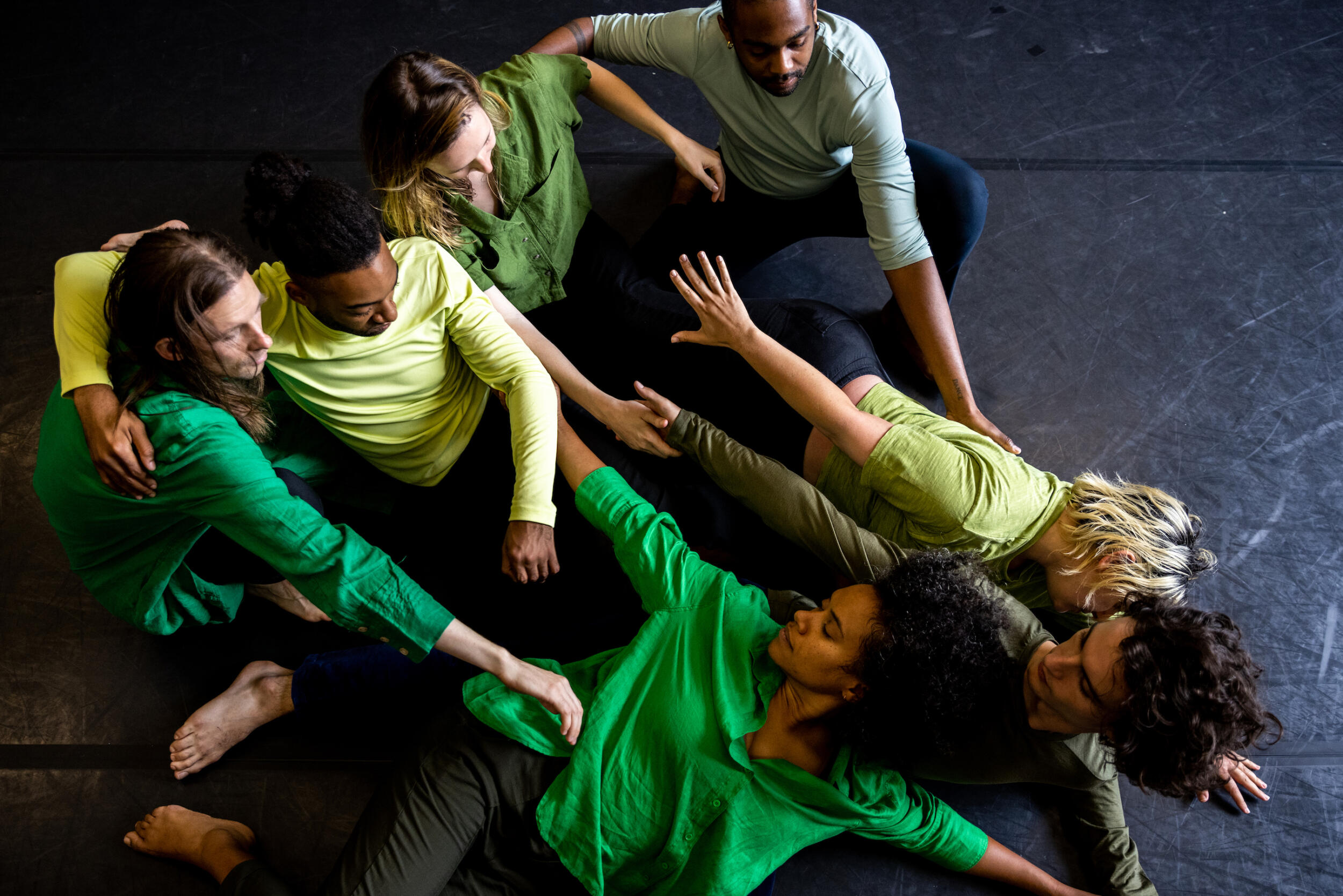 A photograph of a pile of dancers reaching for one another. They wear shirts in varying shades of green.