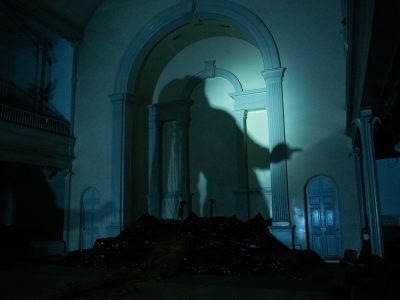 In a darkly lit church, mayfield's larger than life shadow is cast on the arched and embellished walls of the altar. Their head is tilted back and their braids fall behind them. The altar steps are covered in a mysterious, black, shiny material.