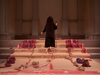 iele paloumpis descends the altar steps backward, with their left arm extended with the palm facing forward, and the right hand holding their white cane. The altar steps are draped with a burlap fabric that has been partially embroidered and there are woven ropes, burlap roses, and other objects adorning the veiled steps.