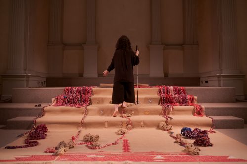 iele paloumpis descends the altar steps backward, with their left arm extended with the palm facing forward, and the right hand holding their white cane. The altar steps are draped with a burlap fabric that has been partially embroidered and there are woven ropes, burlap roses, and other objects adorning the veiled steps.