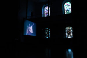In the darkness, light peaks through the stained glass windows of the St. Mark's Church. Glowing in the center of the space is a projection screen with an image of John Bernd performing projected on the screen.