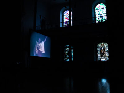In the darkness, light peaks through the stained glass windows of the St. Mark's Church. Glowing in the center of the space is a projection screen with an image of John Bernd performing projected on the screen.