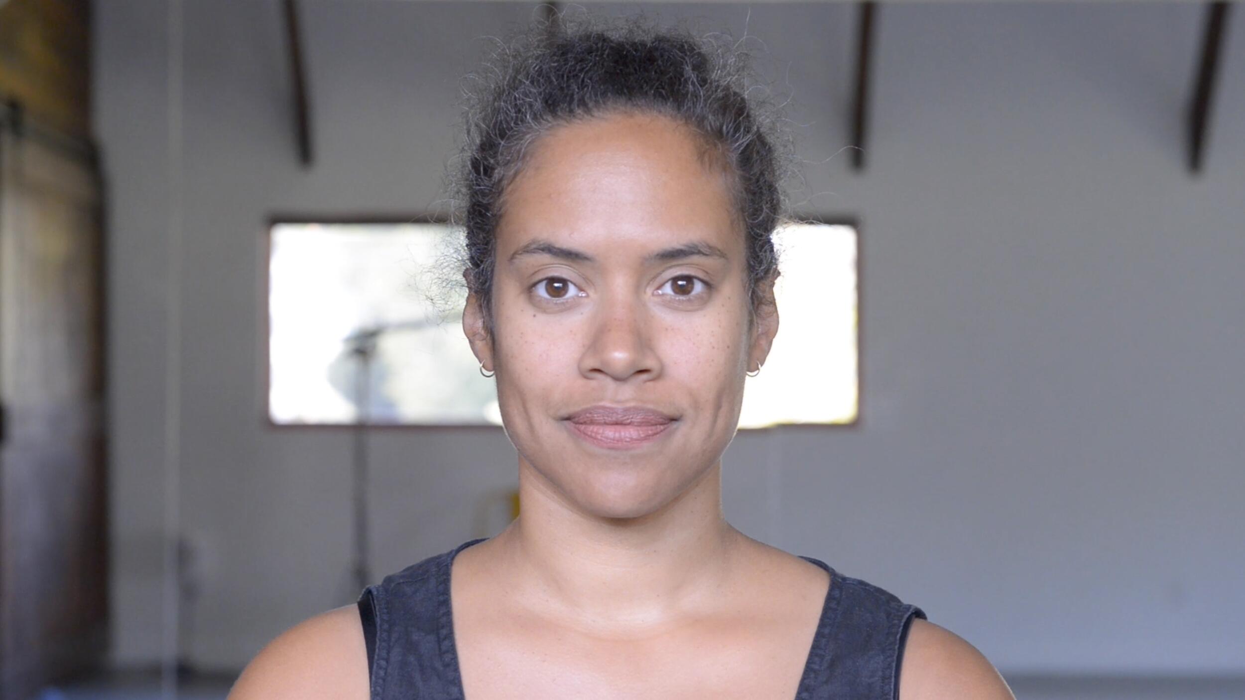 A video still portrait of a light-skinned black woman in a bright loft with salt and pepper disheveled hair pulled back, a slight smile, wearing a black tank top.