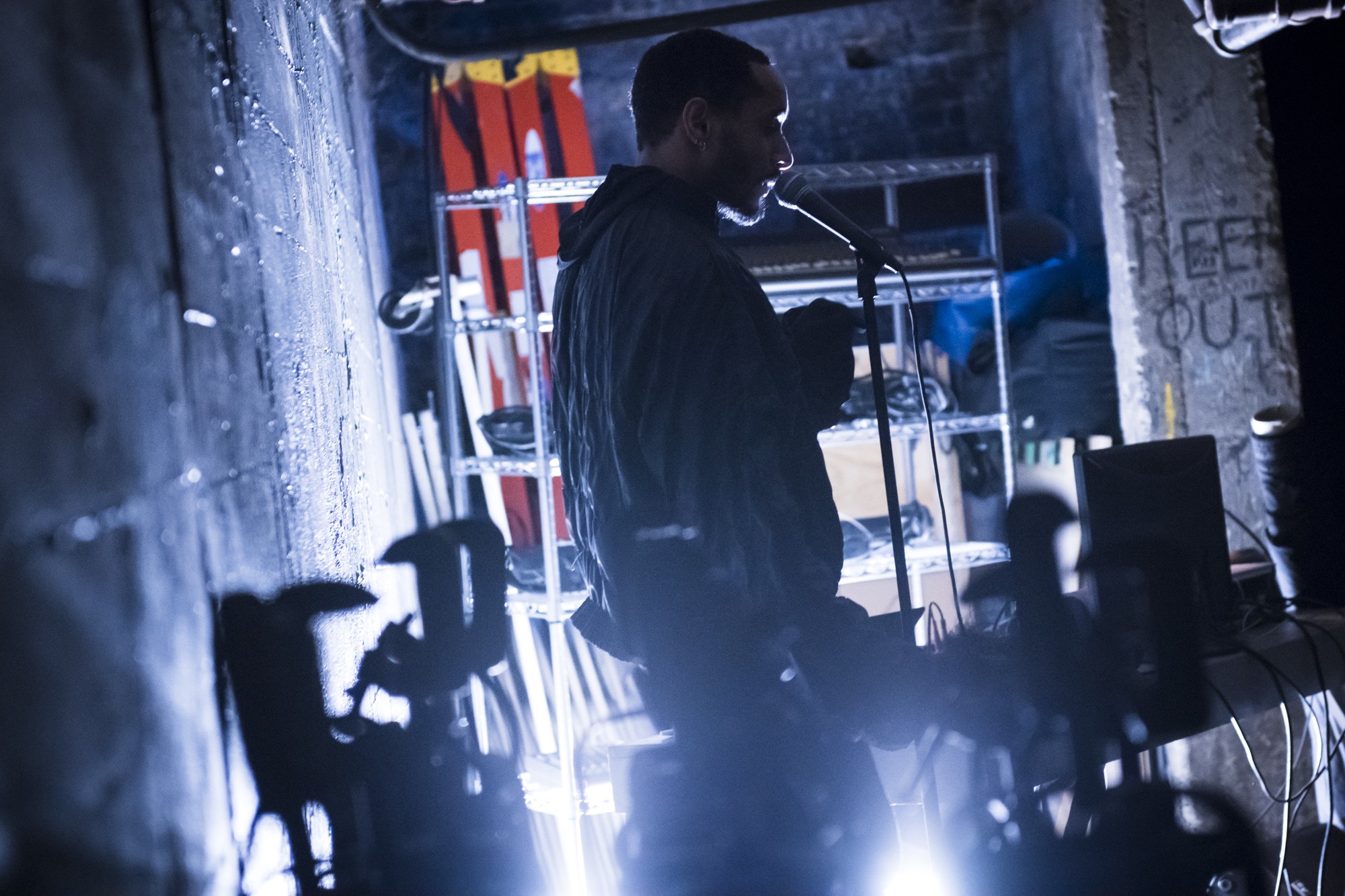 Niall is standing in front of a microphone (very likely speaking or singing) on a makeshift stage and theater tech console in the basement of the Chocolate Factory at its former location on 49th Avenue performing in his 2019 work Fantasies in Low Fade. This photograph was taken by Paula Lobo.