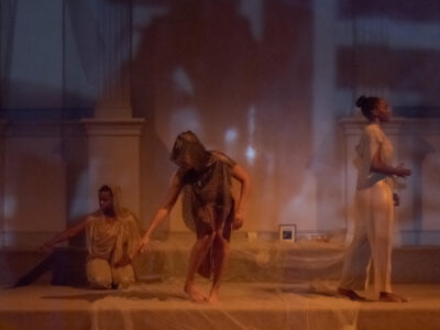 In a hazy and shadowy lighting, three black femme dancers are placed on the altar steps. Leslie is seated on her knees with an arm outstretched to the ground. Paloma, in the middle, is standing crouched, also with her right arm outstretched toward the ground. Tenisha walks facing away from the group.